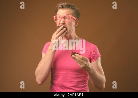 Studio shot von jungen hübschen Nerd mann Essen Schokolade Kuchen so unordentlich Konzept gegen die braunen Hintergrund Stockfoto