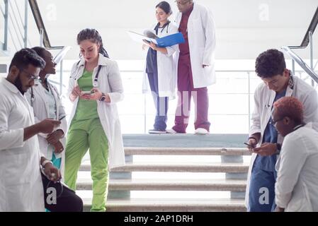 Junge Ärzte, gemischten Rennen, Sitzen, Stehen auf der Treppe, im Flur des Krankenhauses. Sie betrachten x-Strahlen, die Verwendung von Minianwendungen in der Medizin zu arbeiten. Peo Stockfoto