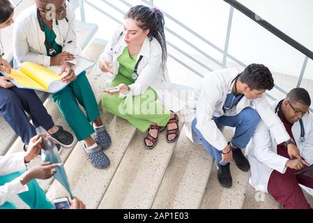 Junge Ärzte, gemischten Rennen, Sitzen, Stehen auf der Treppe, im Flur des Krankenhauses. Sie betrachten x-Strahlen, die Verwendung von Minianwendungen in der Medizin zu arbeiten. Peo Stockfoto