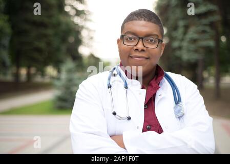 Porträt einer jungen afrikanischen Arzt. Ein Mann in einem weißen Mantel, eine chirurgische Anzug, mit einem Stethoskop, in Gläser, lächelt. Im Freien Stockfoto