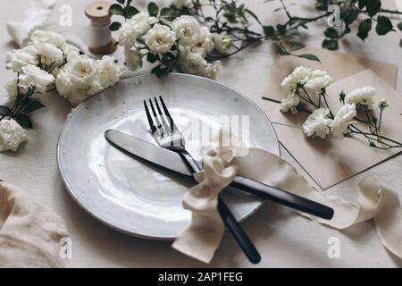 Nahaufnahme von festlich gedeckten Tisch mit schwarzem Besteck, weiße rose Blumen, Seidenbändern, Porzellan, Teller und Briefumschläge auf weißem Hintergrund. Stockfoto