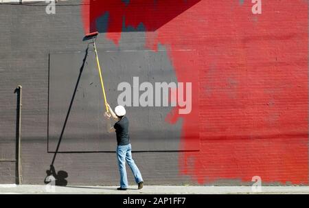 Handwerker in der Mitte durch die Malerei eine graue Wand mit roter Farbe mit einer langen Stange und einer Rolle Stockfoto