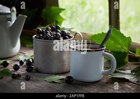 Emaillierte Schale gesunder Kaffee mit Schwarze Johannisbeere Beeren, Becher von schwarzen Johannisbeeren, Kaffee Wasserkocher auf Holztisch. Stockfoto