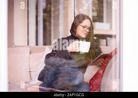 Der hübsche junge Frau sitzt hinter Glas Fenster im Wohnzimmer, während mit digitalen Tablet und Tee trinken. Stockfoto