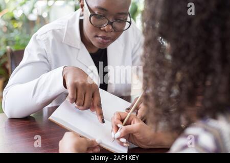 Patienten Mädchen mexikanischen Zeichen eine Behandlung mit einer schwarzen Frau Doktor junge Menschen von gemischten Rennen im Doktorbüro. Gemütliche Atmosphäre Lächeln p Stockfoto