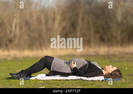 Schwangere Frau ruht auf dem Rücken auf dem Rasen draußen in der Natur an einem sonnigen Tag, Verlegung auf weiche Decke auf dem Boden mit geschlossenen Augen und halten Sie Stockfoto