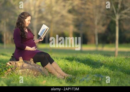 Schwangere Frau mit einem Buch sitzen draußen im Park an einem sonnigen Tag. Volle Länge Seite Porträt mit Kopie Raum Stockfoto