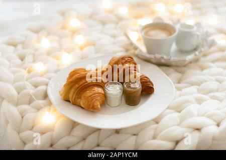 Frische Croissants mit Marmeladen und Americano mit Milch auf gestrickter weißer Wolldecke und leuchtenden Girlanden. Gemütlicher Wintermorgen zu Hause. Skandinavisches Schlafzimmer. Stockfoto