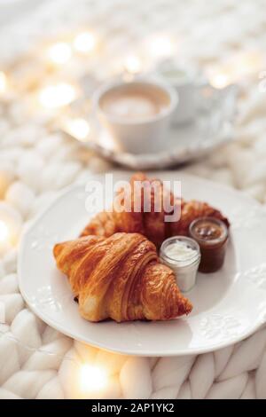 Frische Croissants mit Marmeladen und Americano mit Milch auf gestrickter weißer Wolldecke und leuchtenden Girlanden. Gemütlicher Wintermorgen zu Hause. Skandinavisches Schlafzimmer. Stockfoto