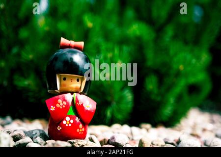 Japanische Kokeshi-Puppe aus Holz, in traditionellem Kimono gekleidet, vor Kiefernzweigen stehend, auf dekorativen Steinen im Garten Stockfoto