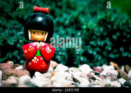 Japanische Kokeshi Puppe aus Holz in traditionellem Kimono, die vor einer Kiefer auf dekorativen Steinen im Garten steht. Handbemalt in rot und Flor Stockfoto