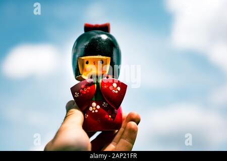 3/4-Hand, die eine japanische Kokeshi-Puppe aus Holz hält, die in traditionellem Kimono über weichem Himmelshintergrund gekleidet ist, handgeschnitzt und in Blumenmustern gemalt ist Stockfoto