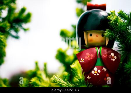 Japanische Kokeshi-Puppe aus Holz, in traditionellem Kimono gekleidet, versteckt zwischen Kiefernzweigen, handbemalt in Rot und Blumen Stockfoto