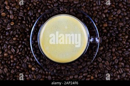 Hohen winkel Nahaufnahme auf transparenten Kaffee Tasse griff auf Glas Untertasse mit Cafe Crema auf unzähligen gerösteten Bohnen verschwommenen Hintergrund Stockfoto