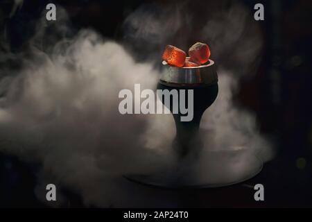 Trendige Hookah-Schüssel mit roten heißen Kohlen in Wolken von schönem weißem Rauch auf dunkelschwarzem Hintergrund. Moderne Shisha mit Kokoskohle für Entspannung und Stockfoto