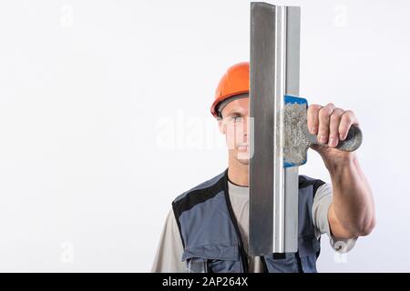 Die Builder verbirgt sein Gesicht hinter einem Spachtel. In Arbeitskleidung und harten Hut. Auf einem hellgrauen Hintergrund. Stockfoto