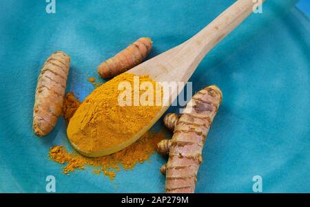 Nahaufnahme von isolierten Holz Löffel mit gelber Curcuma Puder und Kurkuma Wurzeln auf Blau alte verkratzte China Teller Stockfoto
