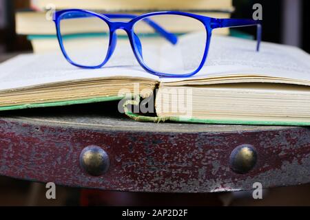 Blick auf alte open vergilbte Buch mit Blau Brille auf antiken Holztisch mit unscharfen Stapel Bücher Hintergrund Stockfoto