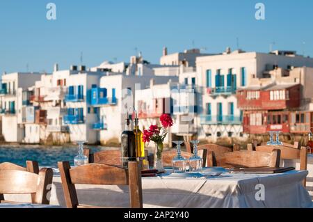 Helle, malerische Nachmittagssicht auf einen leeren Tisch, der auf einen Sonnenuntergang wartet, der am Hafen in der griechischen Altstadt von Mykonos eingerichtet wurde Stockfoto