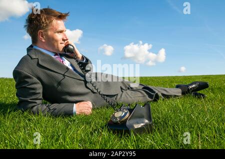 Lächelnd Geschäftsmann auf einem sonnigen Flecken von hellem grün Gras sprechen auf eine altmodische Telefon Stockfoto