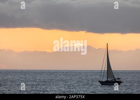 Ein einsames Segelboot, das während eines warmen Sonnenuntergangs auf Gran Canaria über ruhiges Atlantikwasser gleitet, wo goldene Wolken den friedlichen Horizont der Küste erleuchten. Stockfoto