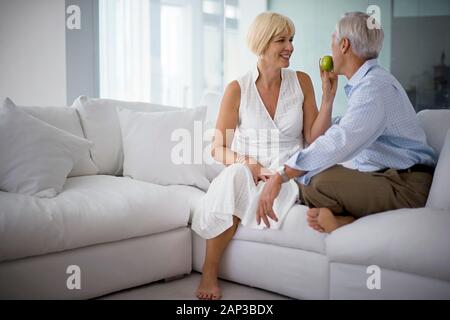 Lächelnd Mitte - erwachsene Frau ihren Ehemann eine Apple sitzen auf einer Couch im Wohnzimmer einziehen. Stockfoto