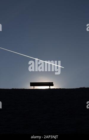 Silhouette einer leeren Bank auf einem Hügel mit Flugzeug- und Kondensstreifen in den klaren, blauen Himmel fliegen von links nach rechts Stockfoto