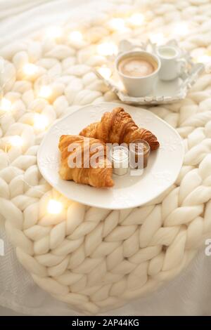 Frische Croissants mit Marmeladen und Americano mit Milch auf gestrickter weißer Wolldecke und leuchtenden Girlanden. Gemütlicher Wintermorgen zu Hause. Skandinavisches Schlafzimmer. Stockfoto