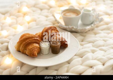 Frische Croissants mit Marmeladen und Americano mit Milch auf gestrickter weißer Wolldecke und leuchtenden Girlanden. Gemütlicher Wintermorgen zu Hause. Skandinavisches Schlafzimmer. Stockfoto