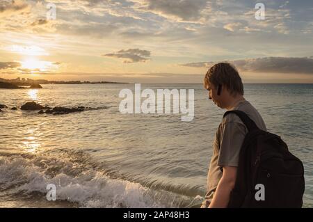 Der kaukasische Mann mit Rucksack geht bei Sonnenuntergang am Hamdeok Beach entlang des Meeres Stockfoto