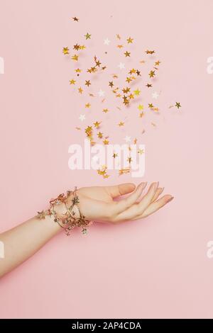 Feier-, Hochzeits- und Geburtstagskonzept. Goldene Konfettisterne, die in weiblicher Hand auf pastellrosa Hintergrund aufblasen Stockfoto