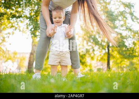 Fröhlicher lächelnder Kinderjunge macht ersten Schritt im Park, Mutter hilft Stockfoto