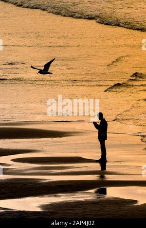 Ein Mann stand auf der Küstenlinie von der intensiven goldenen Licht der untergehenden Sonne auf den Fistral Beach in Newquay in Cornwall. Stockfoto