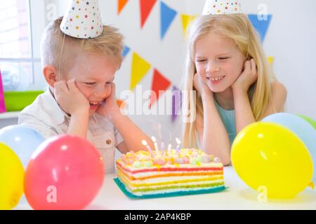 Zwei blonde kaukasische Kinder Junge und Mädchen, die Geburtstags-Regenbogenkuchen mit brennenden Kerzen und Wünsche auf der Geburtstagsfeier betrachten. Farbenfroher Hintergrund Stockfoto