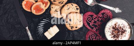 Banner mit Romantischem Frühstück. Zwei Kaffeetups, Cappuccino mit Schokoladeplätzchen und Keksen in der Nähe roter Herzen auf schwarzem Tischhintergrund. Valentine Stockfoto