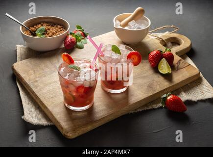 Erdbeere Caipirinha, Kalk, frische Minze, brauner Zucker und zerkleinertes Eis. Rustikale Umgebung mit schwarzem Hintergrund Stockfoto