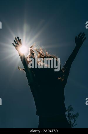 Silhouette der jungen Frau, die ihre Hände mit Freude zur Sonne hebt. Dunkles und schwaches Bild eines glücklichen, nicht erkennbaren Mädchens Stockfoto