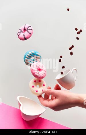 Konzept zum Valentinstag, Levitation von Douglasien über der herzförmigen Schüssel. Kaffeebohnen fliegen in einer Espresso-Tasse. Hand fängt pinkfarbenen Donut. Kreativer Backgr Stockfoto