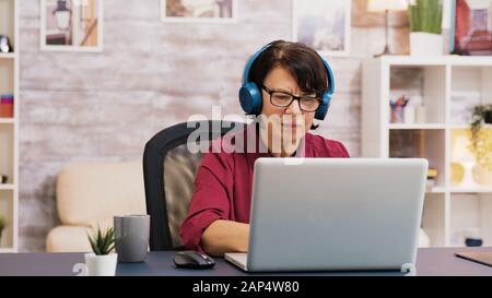 Alte Frau, die eine Tasse Kaffee beim Arbeiten mit einem Laptop mit Kopfhörern auf dem Kopf genießt. Älterer Mann, der im Hintergrund eine Tablette verwendet. Stockfoto
