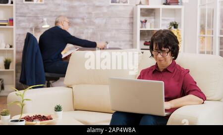 Ältere Frau mit Brille, die auf einem Laptop sitzt und auf dem Sofa sitzt. Älterer Mann liest im Hintergrund ein Buch. Stockfoto