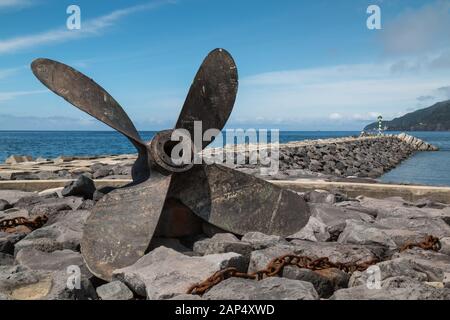 Boot Propeller, im Hafen als Statue verwendet. An der felsigen Küste, im Atlantik im Hintergrund. Blauer Himmel mit weißen Wolken. Povoacao, Sao Miguel, Azoren Stockfoto