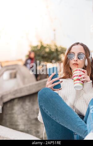 Portrait von schönen jungen trendigen Mädchen mit Kopfhörern und Sonnenbrillen verwendet smart phone Halten sie Fruchtsaft und Sitzen Stockfoto