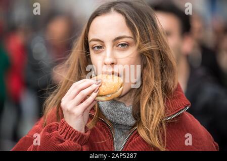 Schöne junge süße Mädchen in modische Kleidung isst fast food Hamburger beim Gehen an einer belebten Straße Stockfoto
