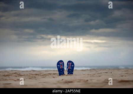 Australische Flaggenthongs, die aufrecht in Sand an einem australischen Strand mit dramatischem Hintergrund von Sturmwolken hängen. Tageskonzept Australien und warten oder beten Stockfoto
