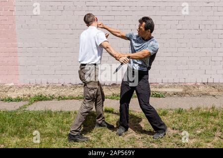 Reifer männlicher Ausbilder schlägt einem Angreifer mit einem Messer ins Gesicht. Straßenkämpfe. Selbstverteidigungsmethoden Krav Maga Stockfoto