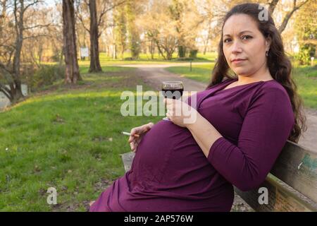 Schwangere Frau mit Alkohol und Zigarette in der Hand, saß auf der Bank im Park und Blick auf Kamera mit fragenden Blick zu. Seite Porträt mit Stockfoto