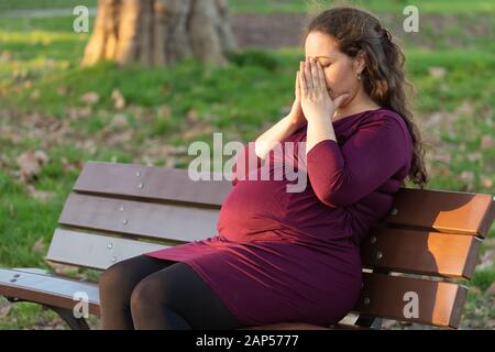 Junge schwangere Frau sich einen Moment Zeit nehmen, zu de-Stress und entspannen Sie sich, wie Sie morgen Krankheit in den warmen Sonnenschein auf einer Parkbank sitzen mit den Händen leidet Cov Stockfoto