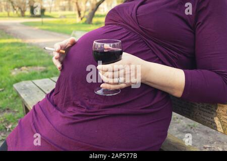 Schwangere Frau auf einer Bank sitzen, trinken Rotwein und Rauchen Zigarette in einem Park. Stockfoto