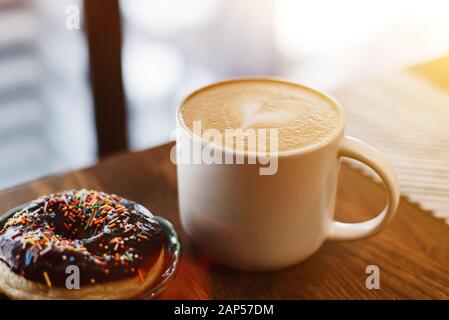 Kaffee mit gezogenem Herz und Milch auf einem Holztisch in einem Café. Schokoladendönut mit Streuung auf dem Tisch neben dem Kaffee. Stockfoto