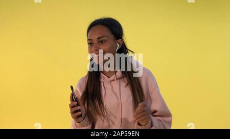 Fröhliches afrikanisches Mädchen, das die Musik in kabellosen Kopfhörern auf gelbem Hintergrund hört. Sie ist melomaniac. Stockfoto